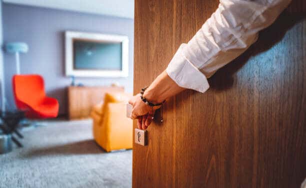 Toned man closing the door of the luxurious hotel room, man wearing a bracelet and a white shirt, closing the wooden door, nice and luxurious hotel room in the background.
