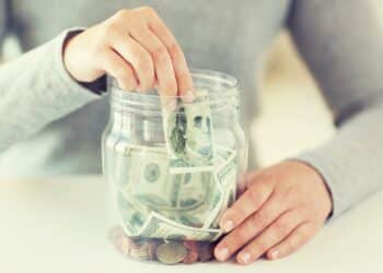 business, finance, saving, banking and people concept - close up of woman hands putting us dollar money into glass mason jar; Shutterstock ID 476500849